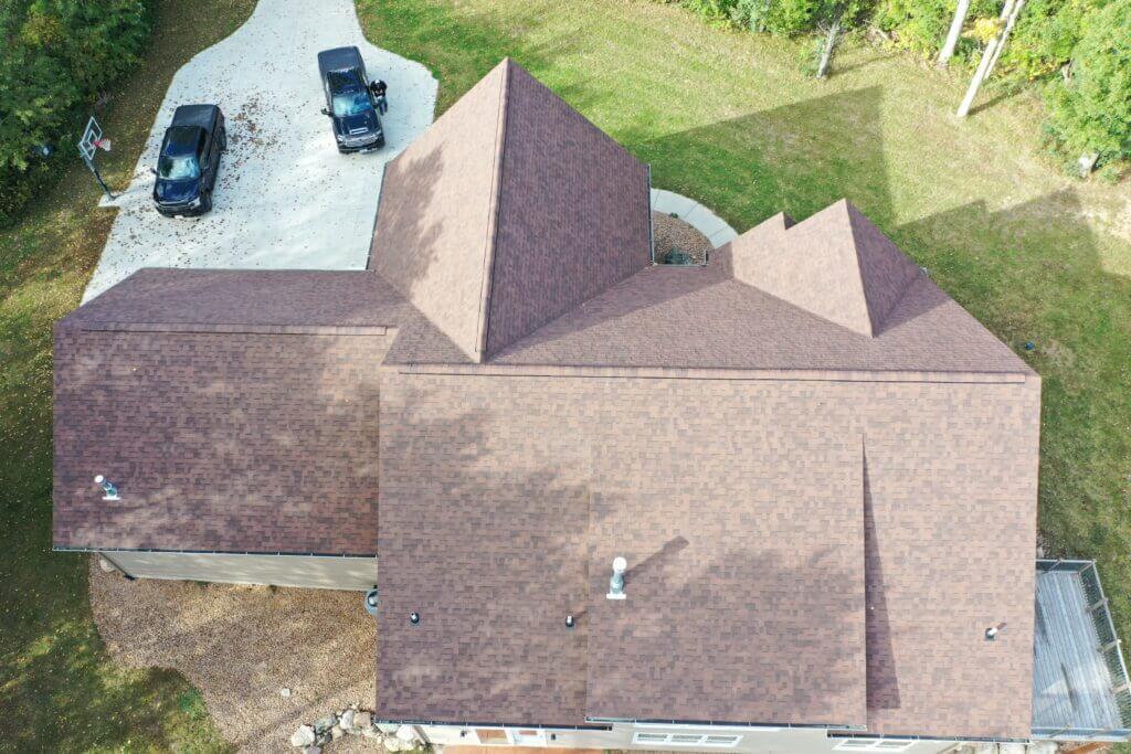 Aerial view of a house with a brown shingle roof, surrounded by grass and trees. Two black vehicles are parked on a light-colored driveway near the house, showcasing quality craftsmanship by Maverick Construction.