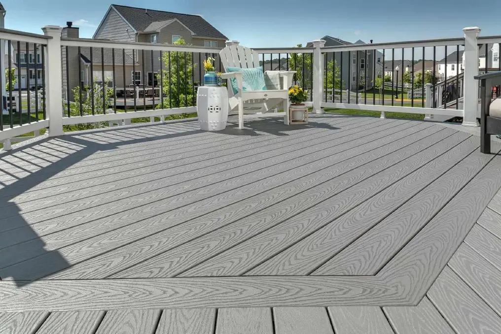 A spacious gray composite deck with white railing showcases a white Adirondack chair, small side table, and potted flowers; suburban houses and greenery are visible in the background.
