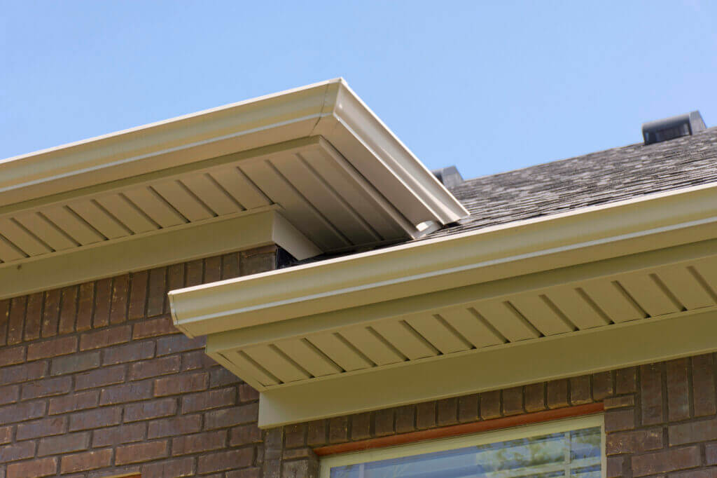 Close-up view of a house’s beige soffit and fascia under the roof eaves, with brown brick walls and a clear blue sky in the background.