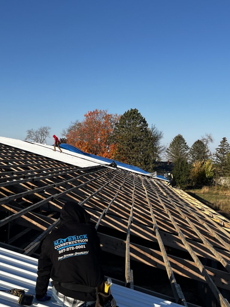 Workers install a metal roof on a large building frame under a clear blue sky, demonstrating expert use of quality roofing materials. One worker in a Maverick Construction hoodie faces away from the camera, while others handle home roofing tasks further up. Trees are in the background.