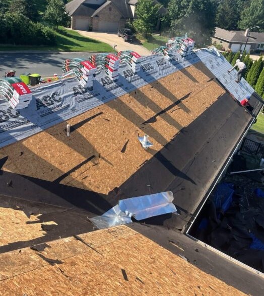 A rooftop under construction with stacks of roofing shingles and exposed wooden panels showcases essential roofing materials. A worker stands near the edge, maintaining safety and efficiency. Houses and trees in the background emphasize the importance of home roofing.